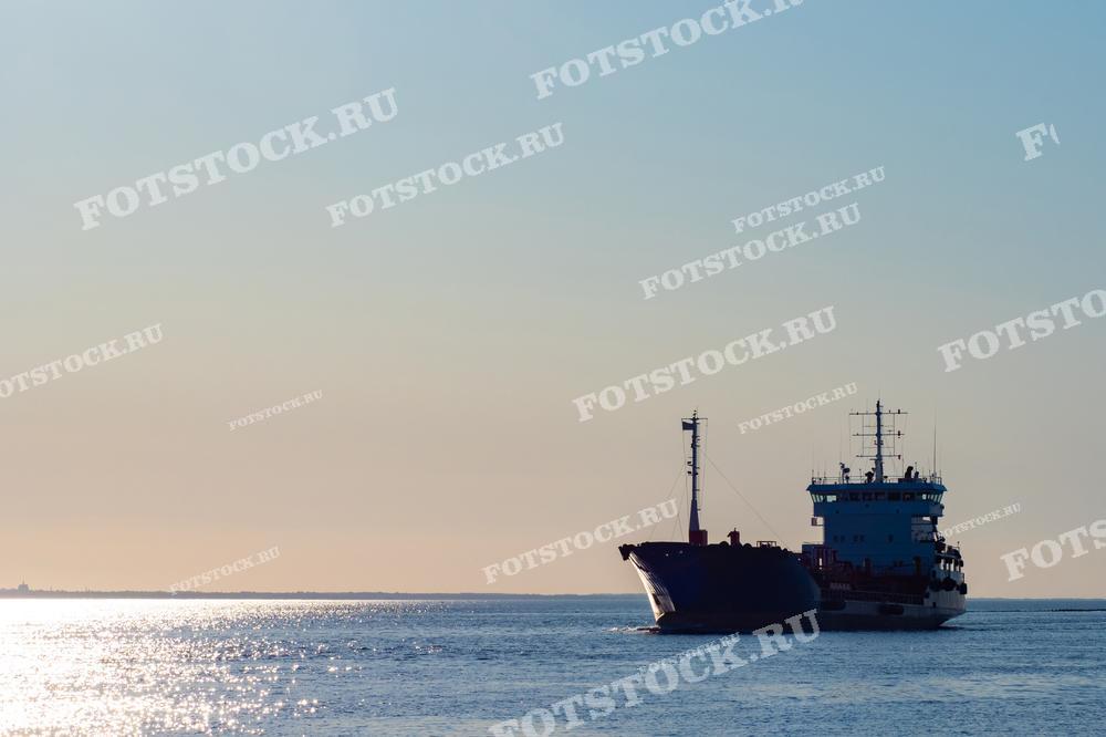 На фотографии изображено грузовое судно, плывущее в открытом море на фоне заката. Вода сверкает в лучах заходящего солнца, создавая живописный вид морской навигации и судоходства.